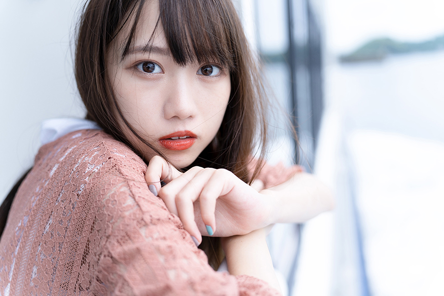 Eine junge Frau mit langen Haaren blickt mit einem rosa Spitzenoberteil in die Kamera. Der Hintergrund ist verschwommen.