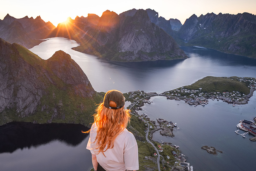 A person with long hair and a cap overlooks a scenic fjord landscape at sunset, with mountains and calm water below.
