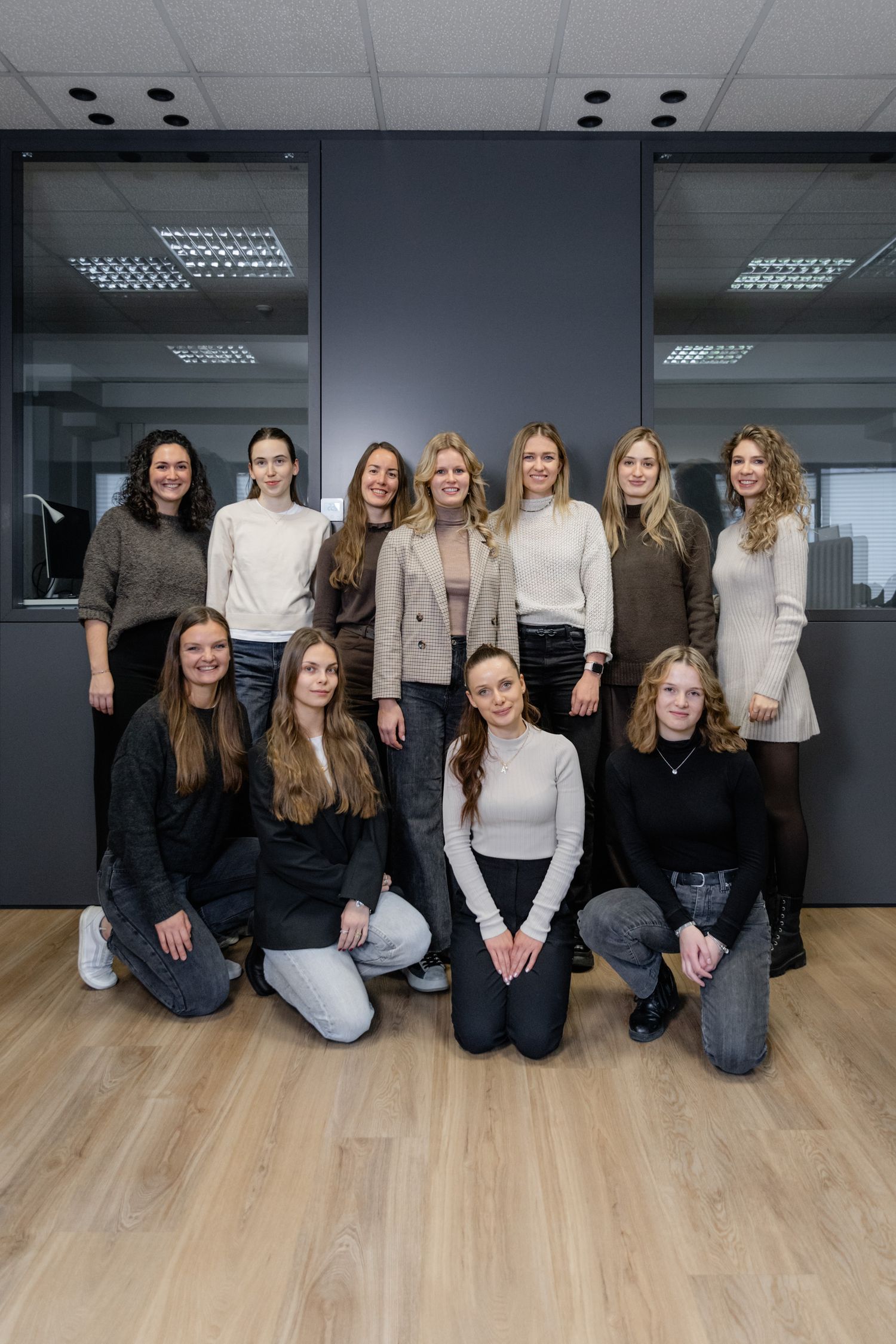 A group of ten women posing indoors in casual and professional attire, standing and crouching on a wooden floor with a dark wall behind them.