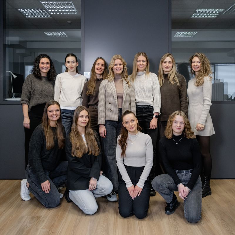 A group of ten women posing indoors in casual and professional attire, standing and crouching on a wooden floor with a dark wall behind them.