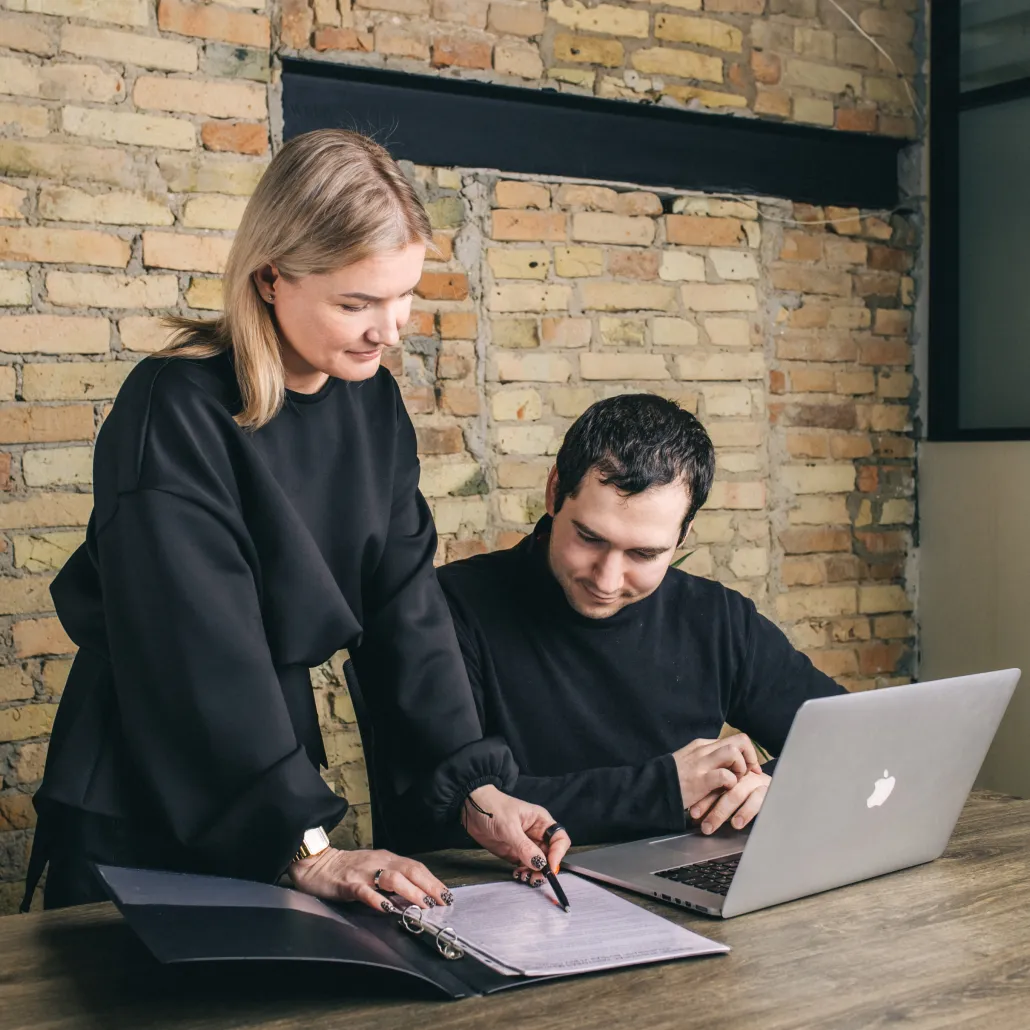 A woman is showing a document to a man with laptop