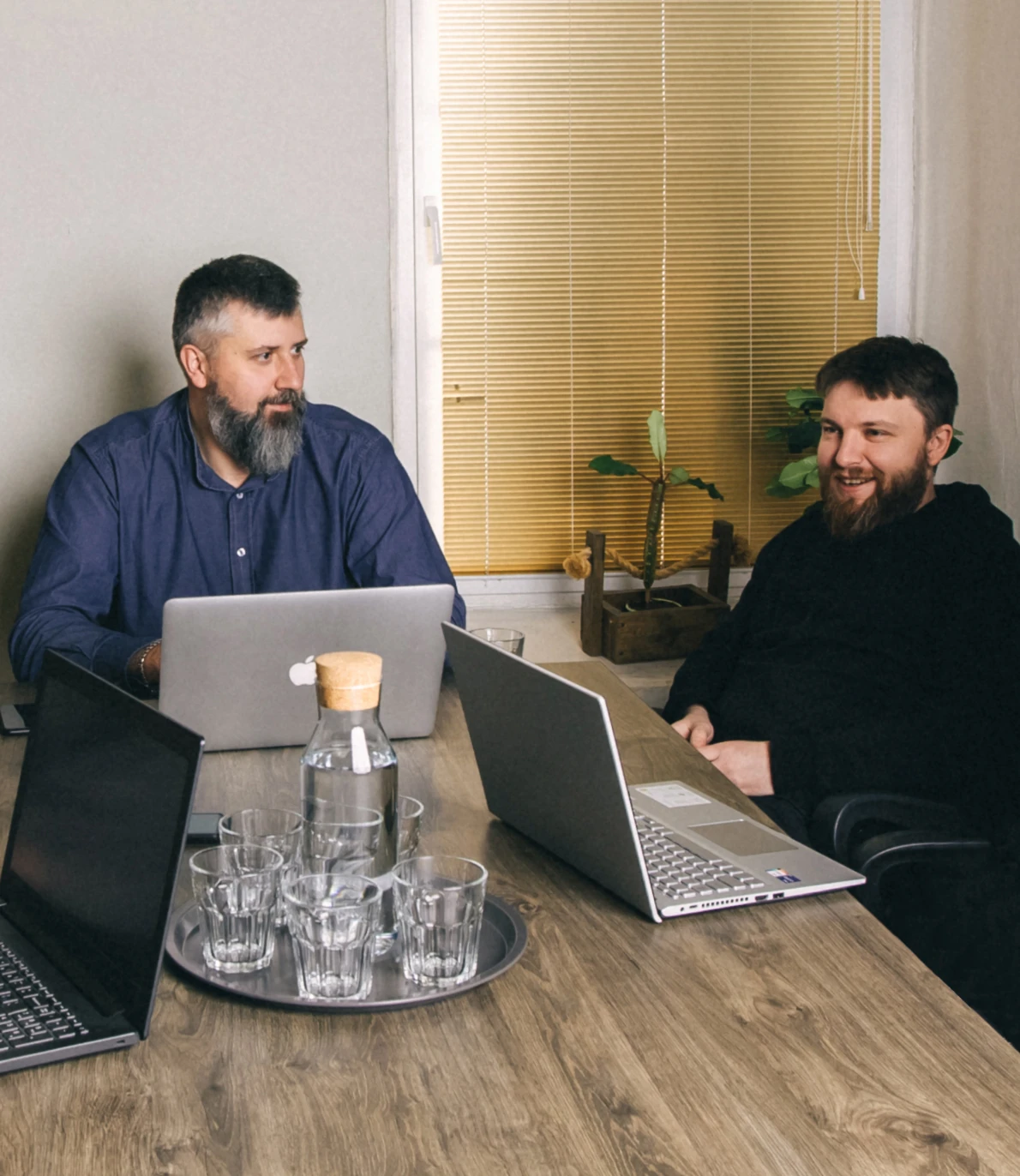 two smiling men are sitting at the table with laptops