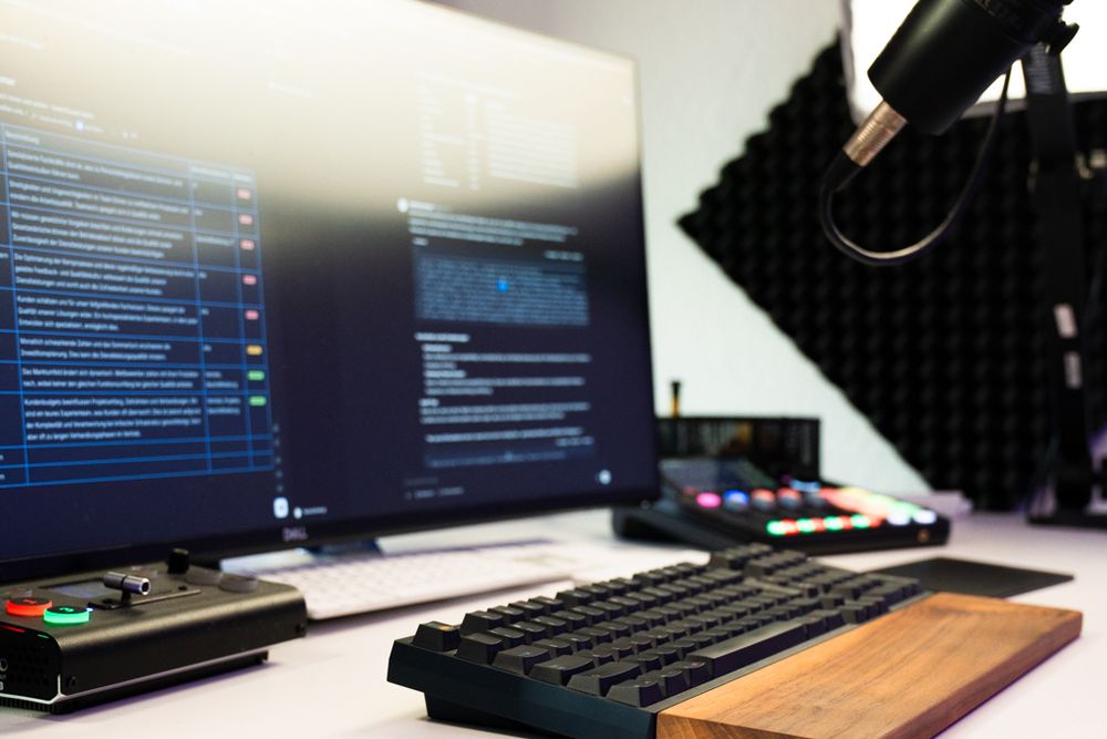 Arbeitsplatz mit zwei Monitoren, Programmcode, Tastatur, Mikrofon und Bedienpult.