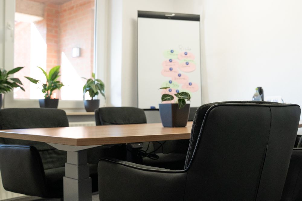 A height-adjustable table with chairs surrounding it. A whiteboard with notes and a bunch of plants are behind them.