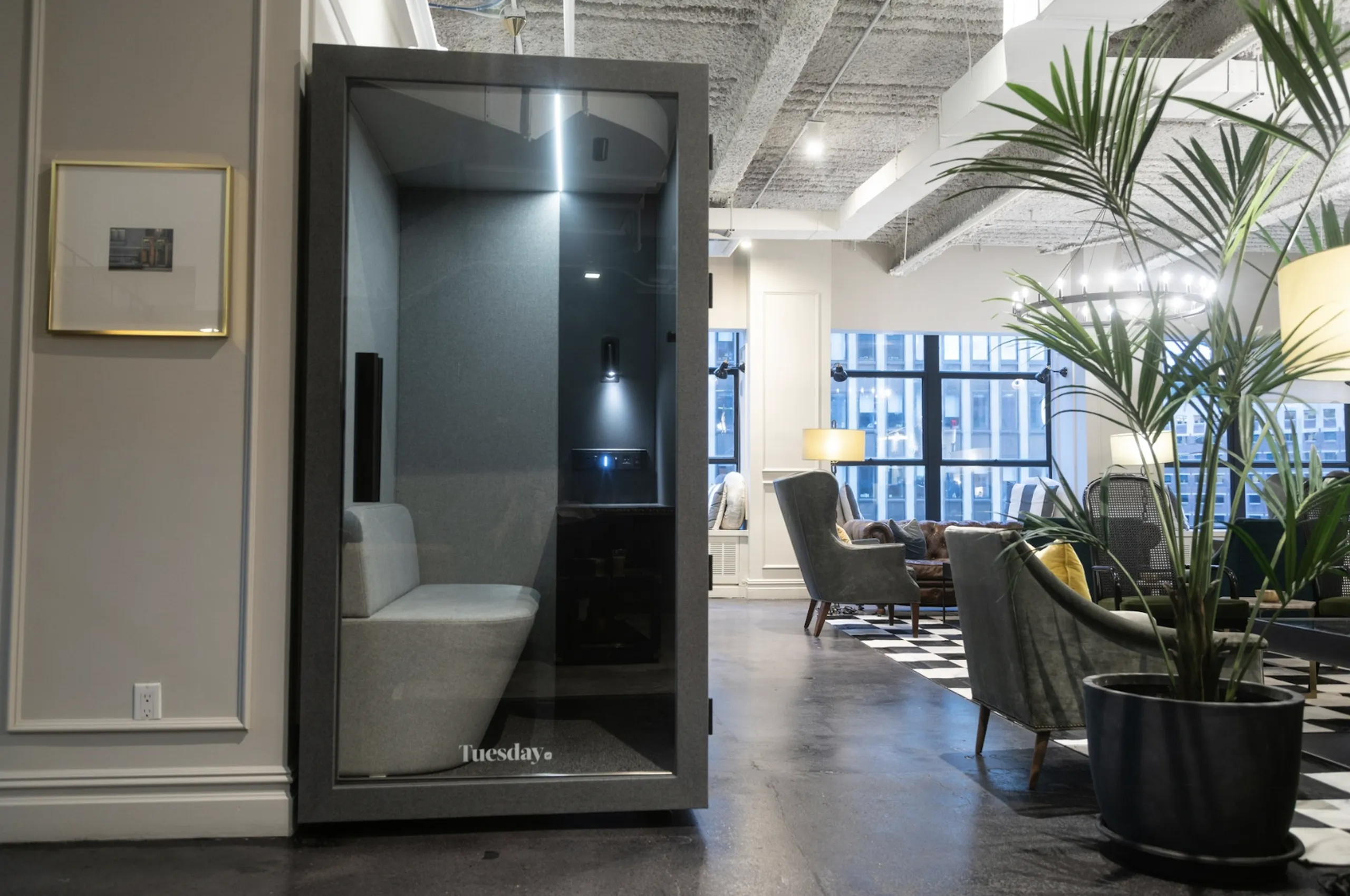 Modern office lounge with a phone booth, gray seating, large windows, and greenery, under exposed ceiling with lights.