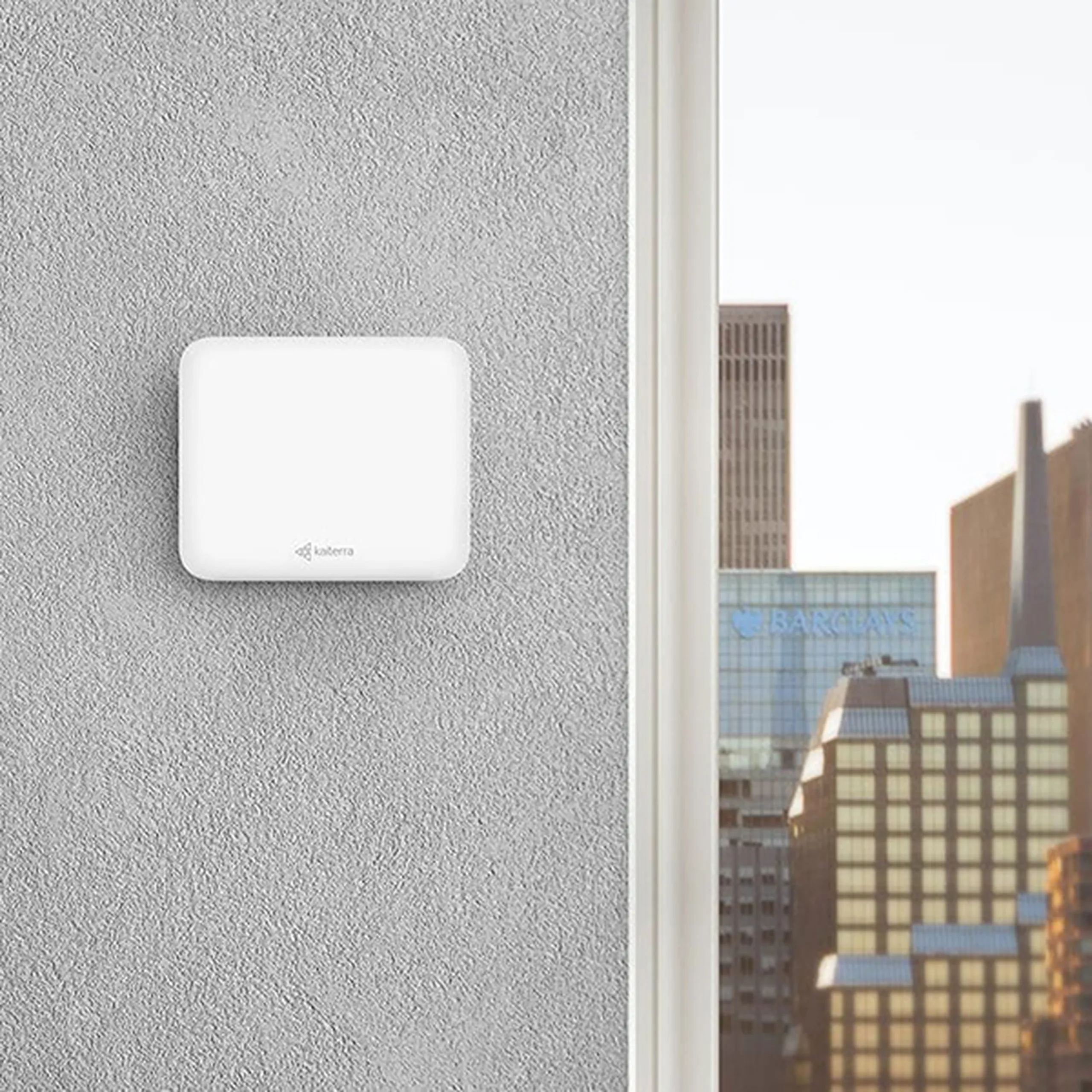 Small white device mounted on a gray wall near a window, overlooking a cityscape with skyscrapers and clear sky.