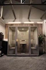 A modern office pod with glass walls, a table, and seating inside. It's centrally located in a room with plants and decorative wood paneling.