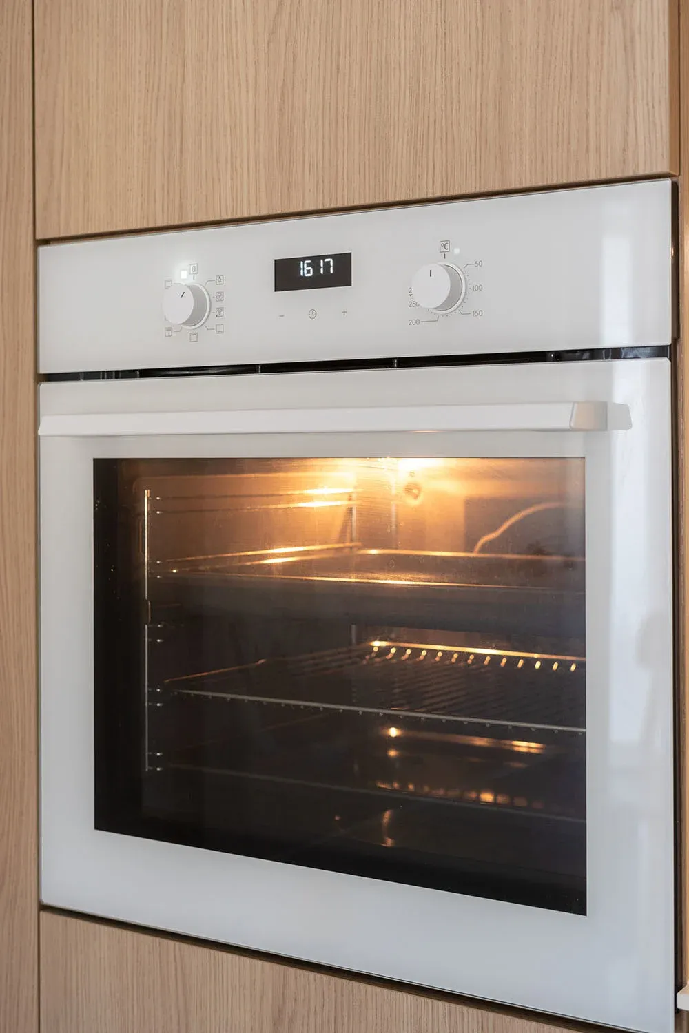 Built-in oven with wood paneling, digital clock showing 16:17, and interior light on, revealing empty racks inside.
