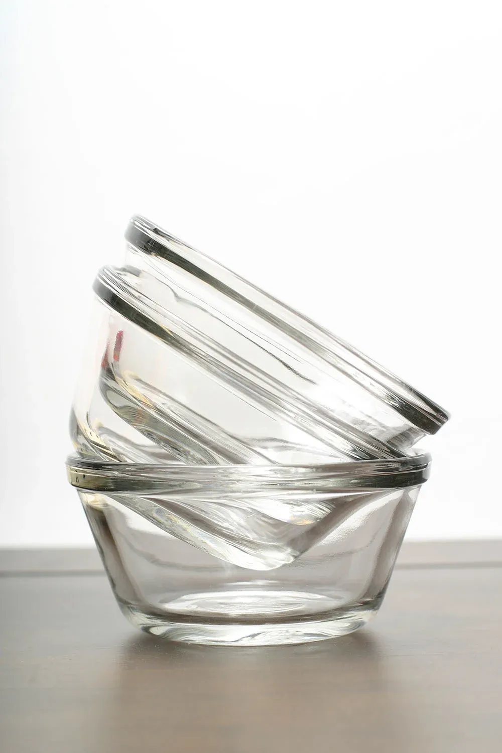 Stacked clear glass bowls tilted on a wooden surface, with a plain white background.