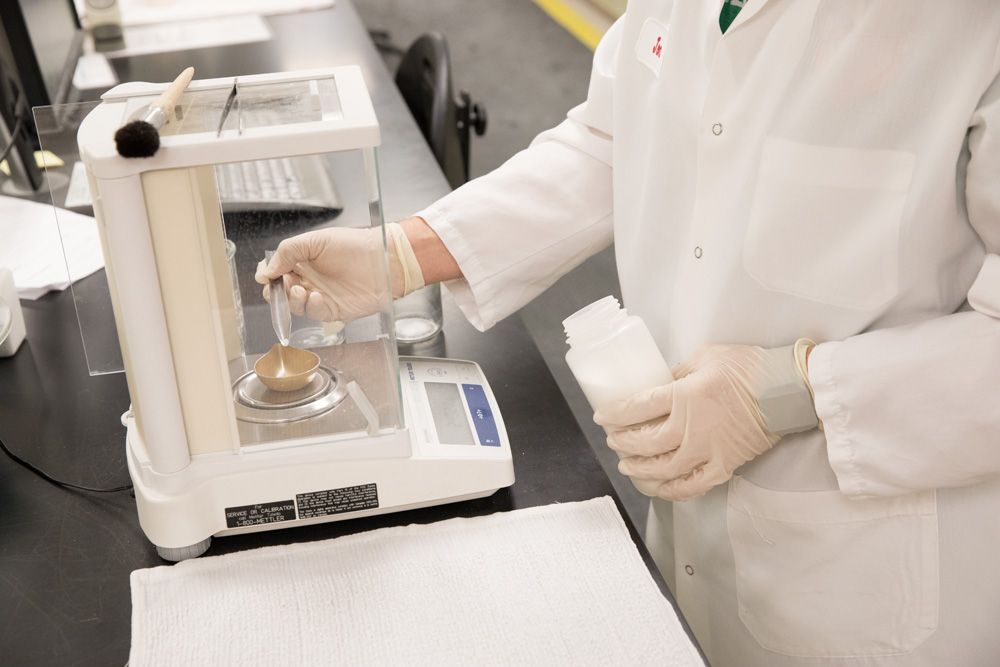 A person in a lab coat and gloves is measuring a substance on an electronic scale while holding a white container in a laboratory setting.