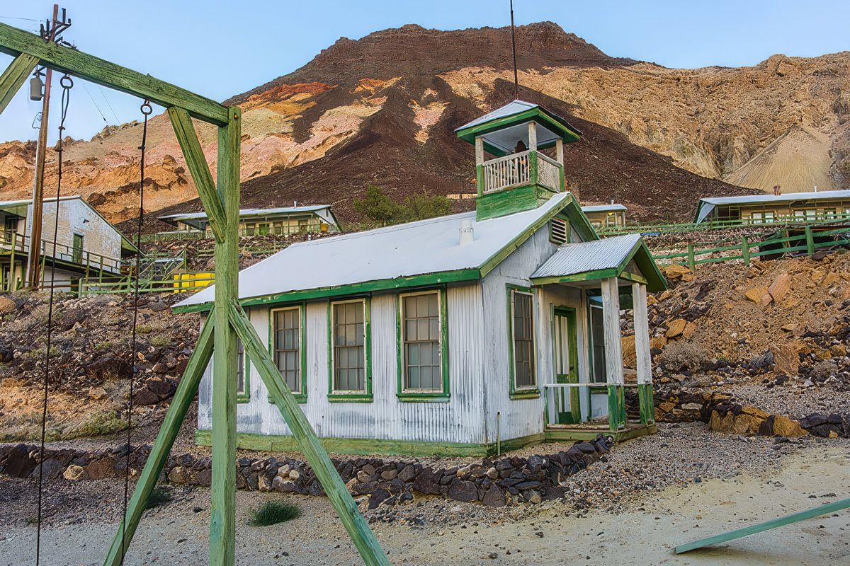 White and green building with a bell tower in a desert landscape, surrounded by rocky hills and swing set in the foreground.