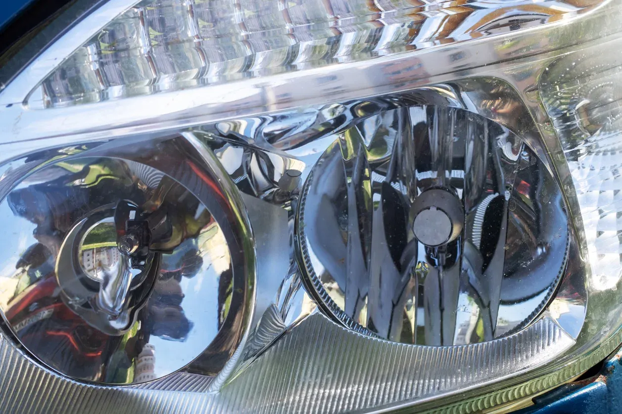 Close-up of a car's reflective headlight assembly with visible reflections of surrounding buildings and sky.