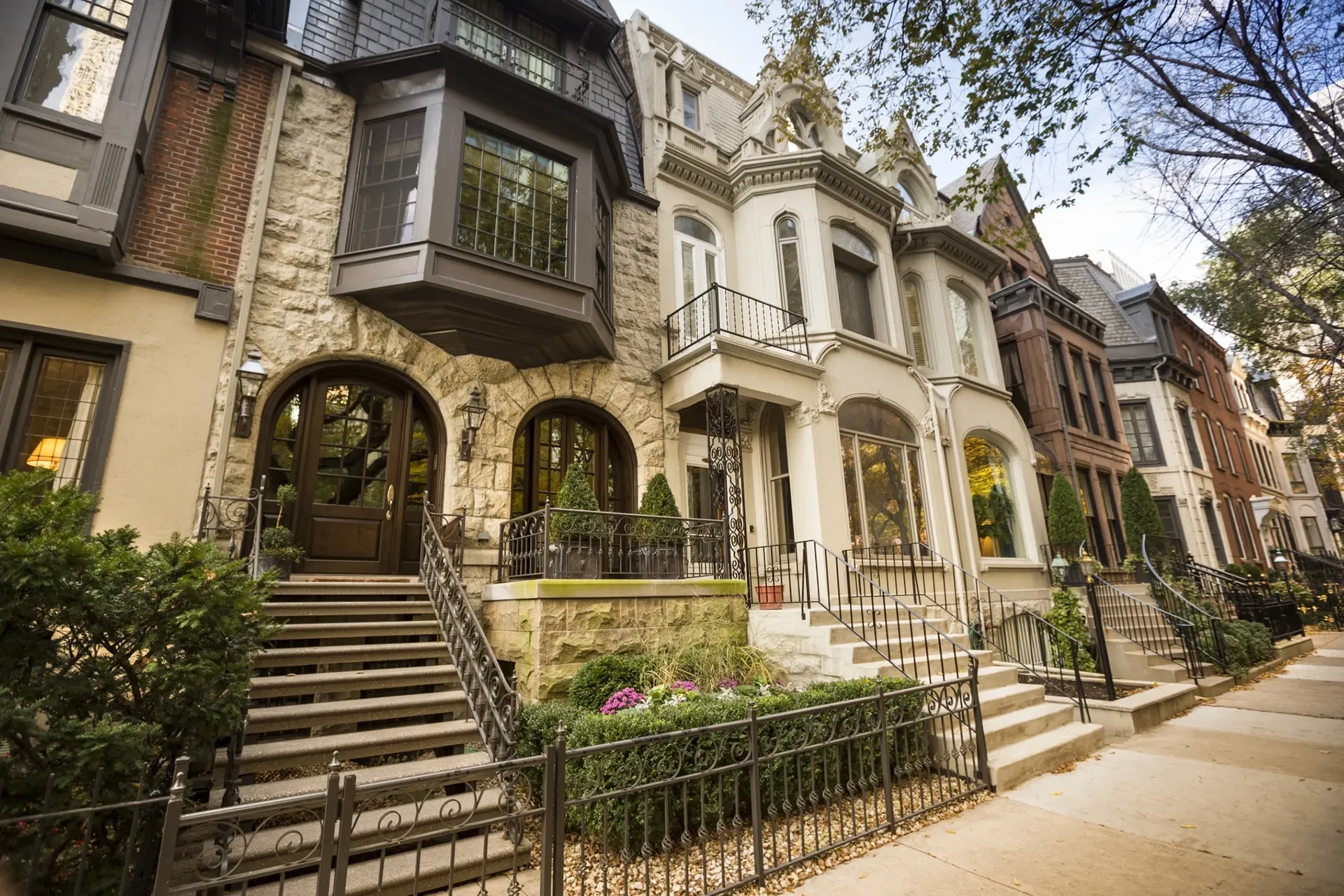 A row of elegant, historic townhouses with ornate facades and decorative railings, bordered by manicured shrubs and trees along the sidewalk.