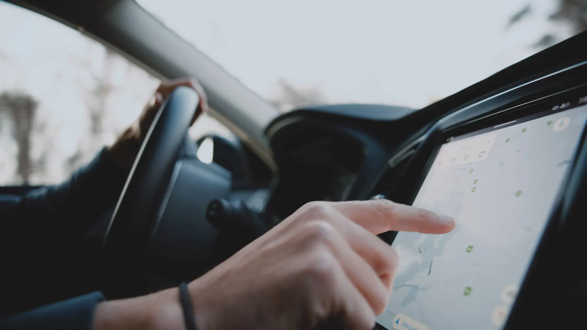 A person in a car is using their finger to navigate a map on a touchscreen GPS device while holding the steering wheel with their other hand.