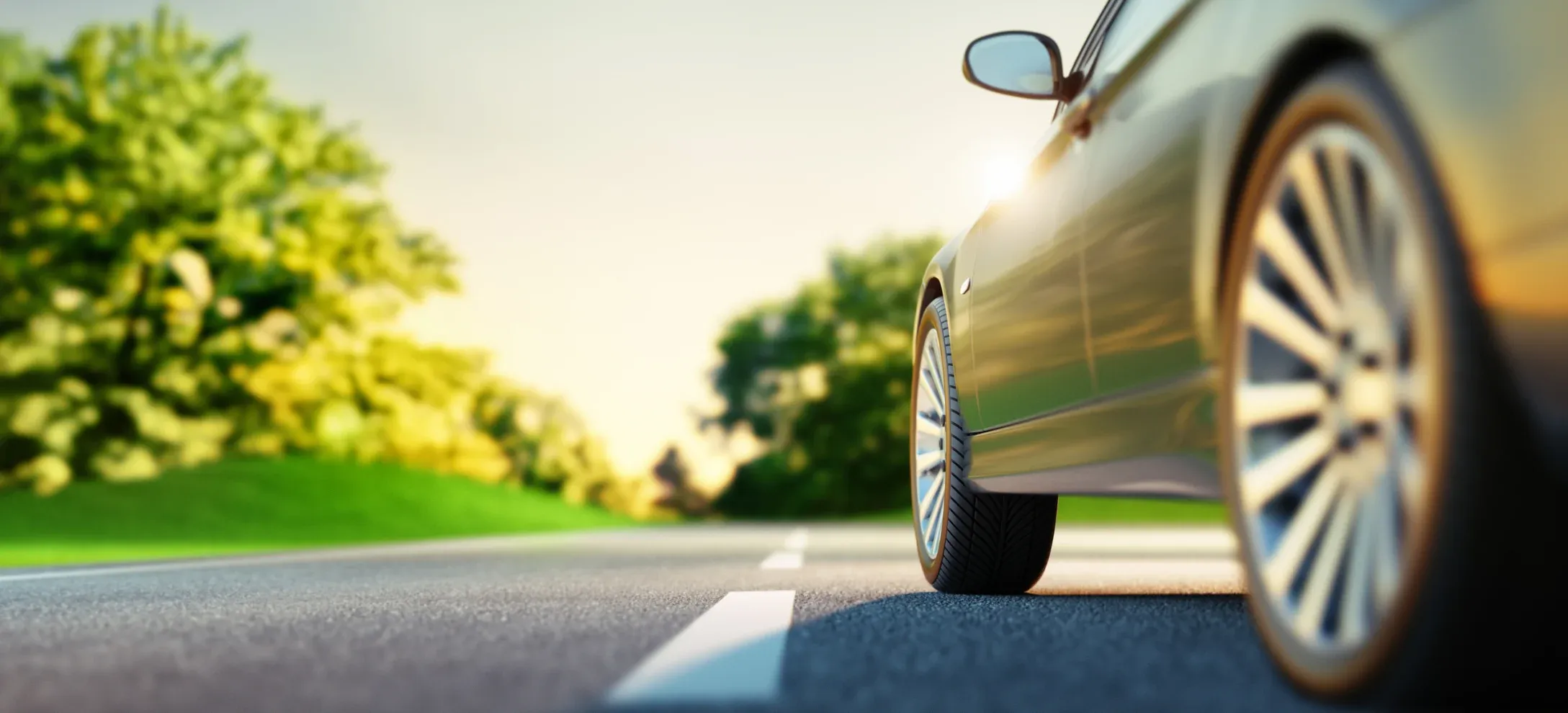 Close-up of a car driving on an empty road with blurred green trees in the background and sunlight reflecting off its side mirror.