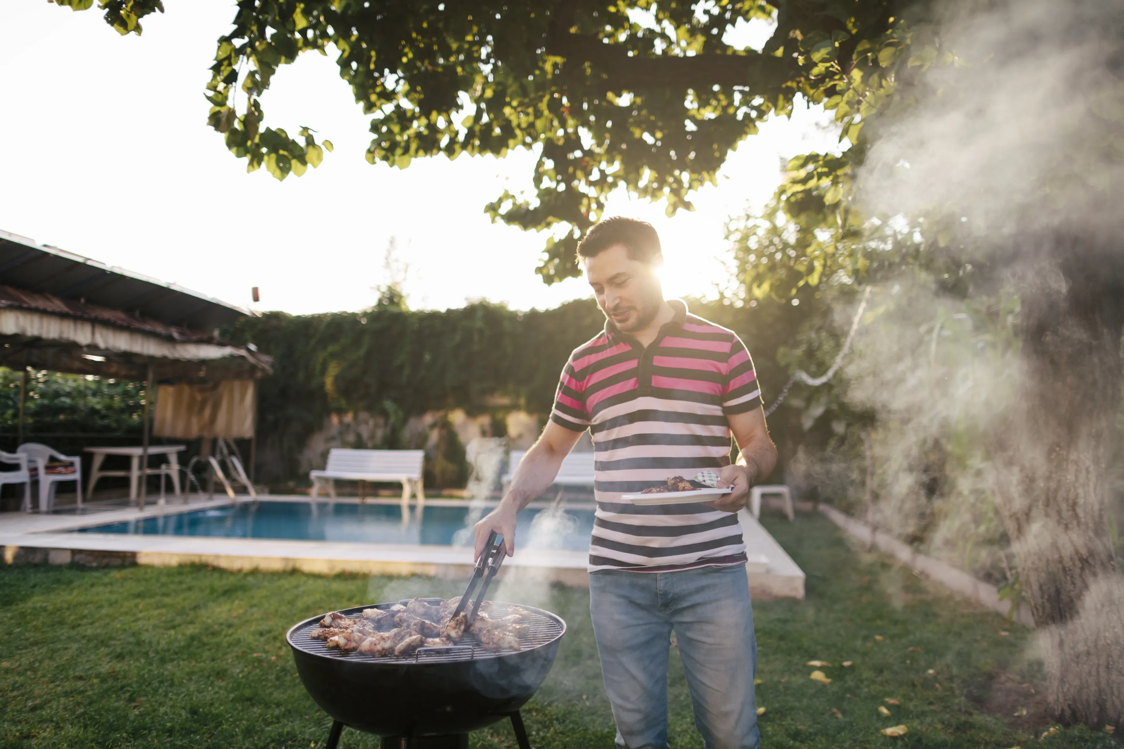 A man grills meat on a barbecue in a sunny backyard with a pool, surrounded by green grass and trees.