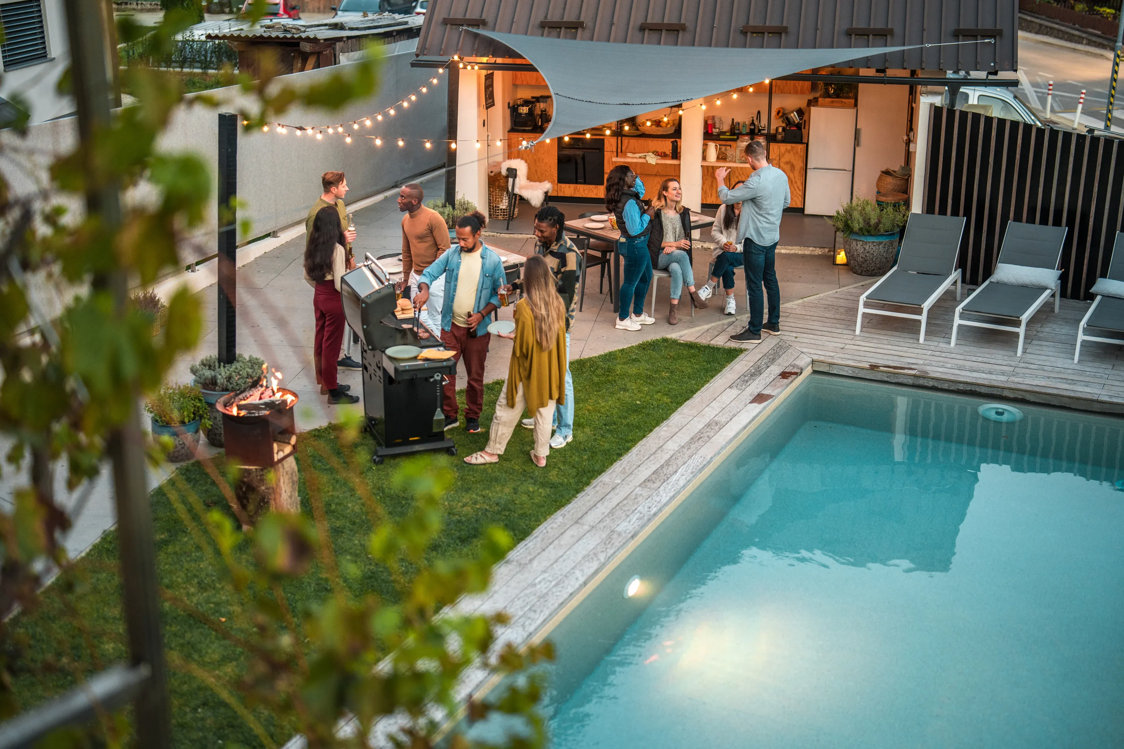 A group of people enjoy a backyard barbecue near a pool, with fairy lights and a cozy outdoor sitting area.