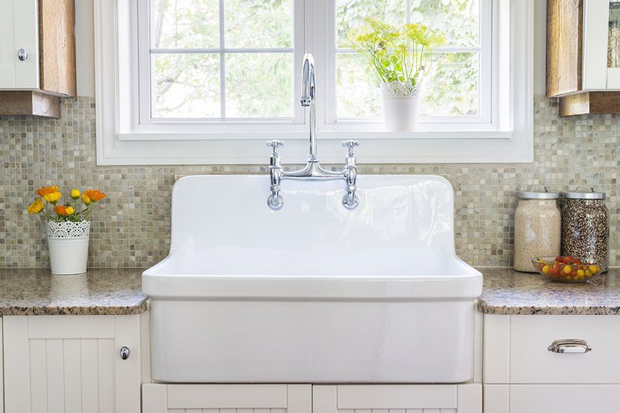 A white farmhouse sink under a window with potted plants on the sill, surrounded by mosaic tile backsplash and jars with food items on the counter.