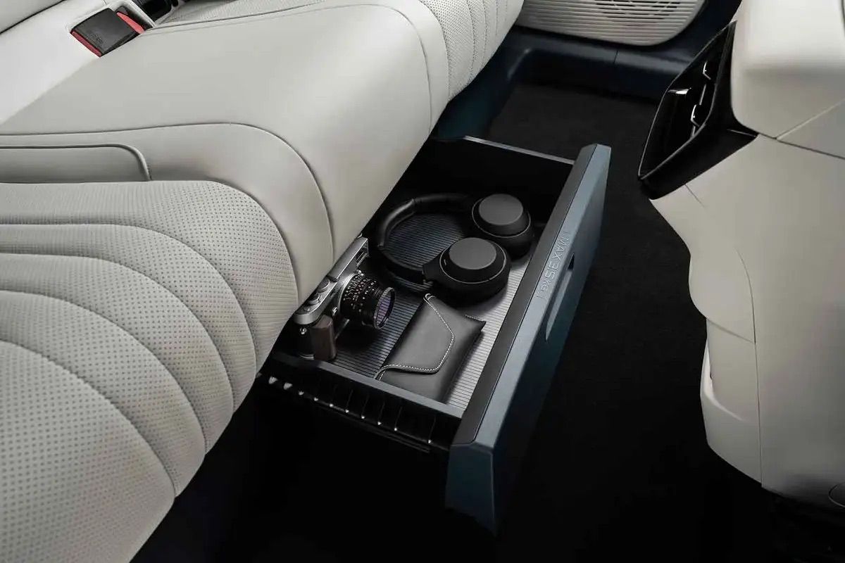 Car interior with a rear seat storage drawer containing headphones, a camera, and a wallet, under light gray perforated leather seats.