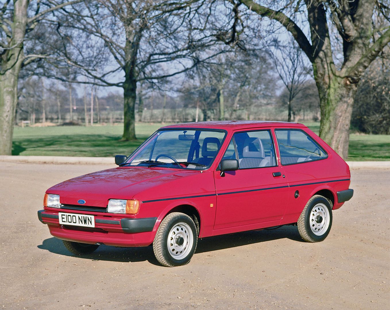 Red hatchback car parked on a dirt road, surrounded by leafless trees in a park setting.