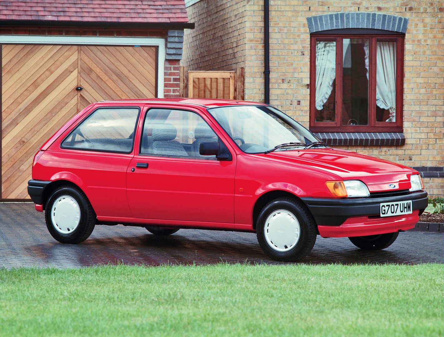 A red hatchback car parked on a brick driveway in front of a house, with windows and a wooden garage door visible in the background.