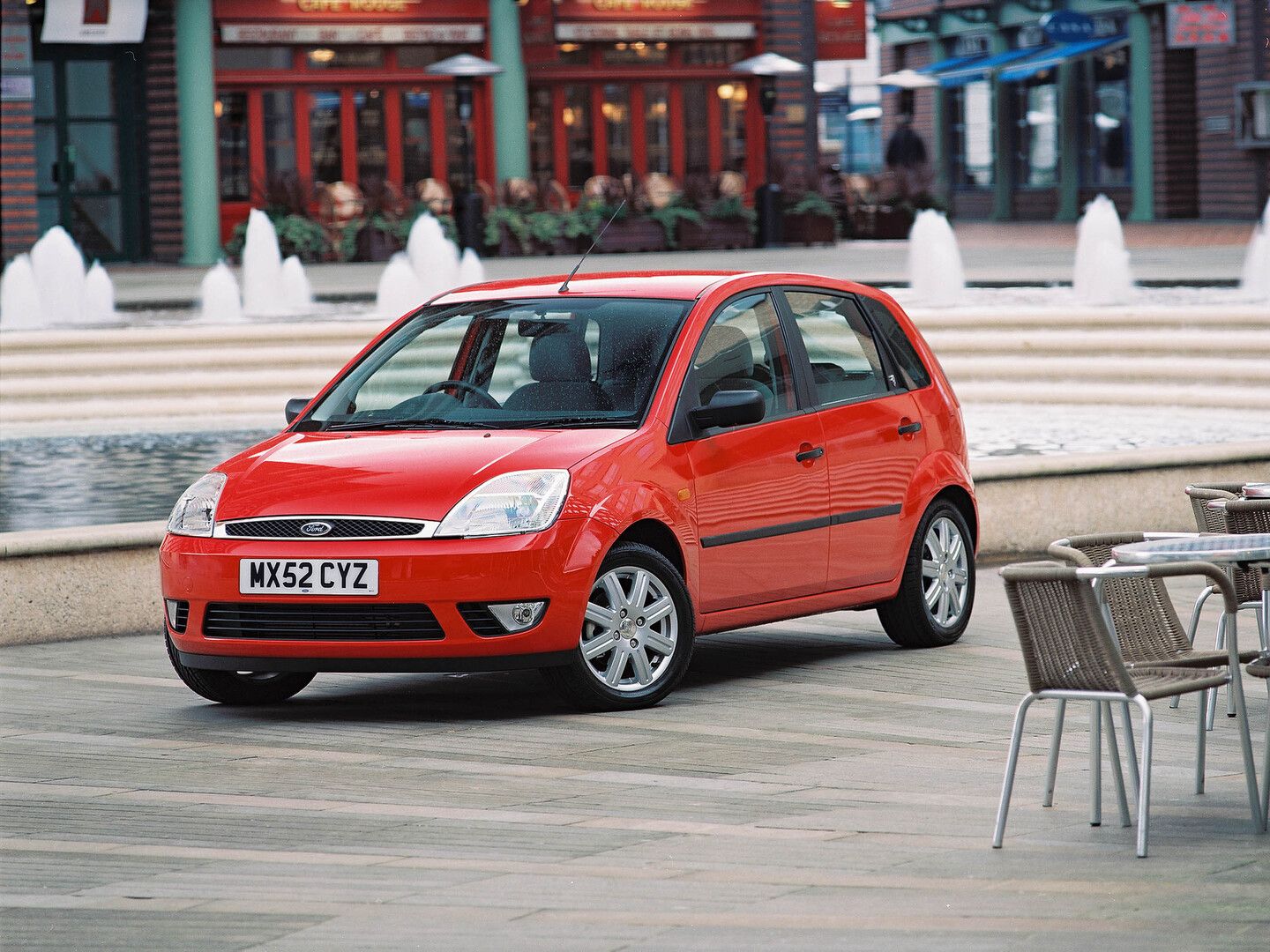 Red Ford hatchback parked on wooden deck near a fountain, with tables and chairs in the foreground and a building in the background.