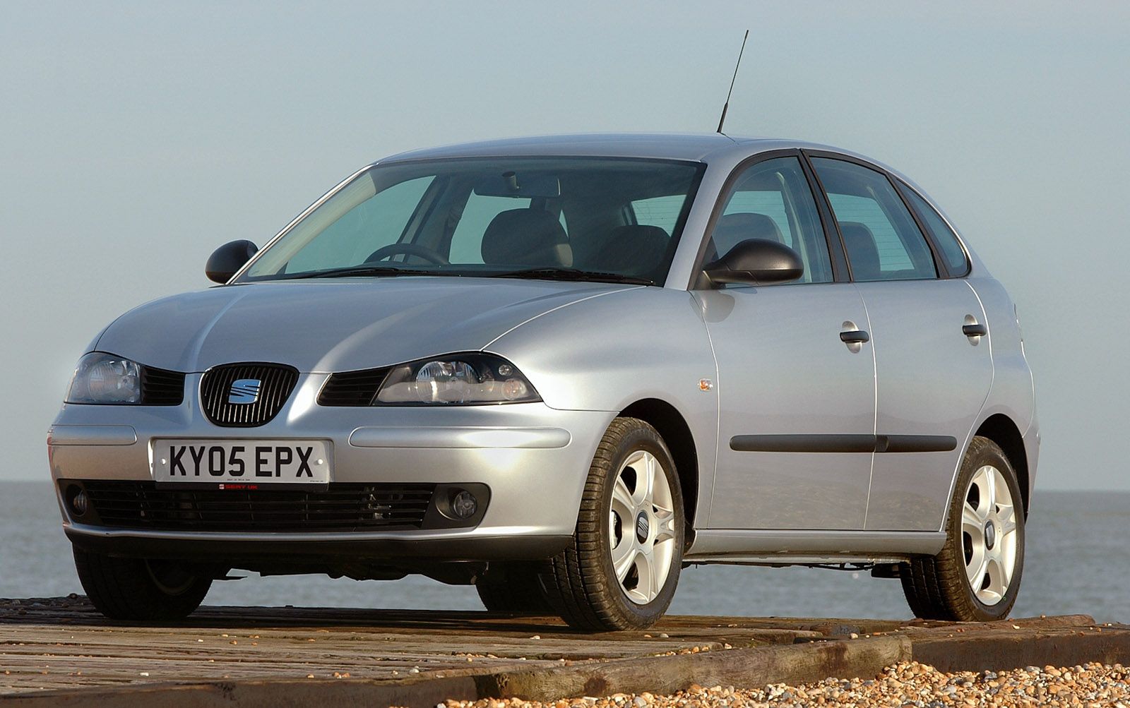 Silver hatchback car parked on a wooden platform by the beach, with a clear sky and calm sea in the background.