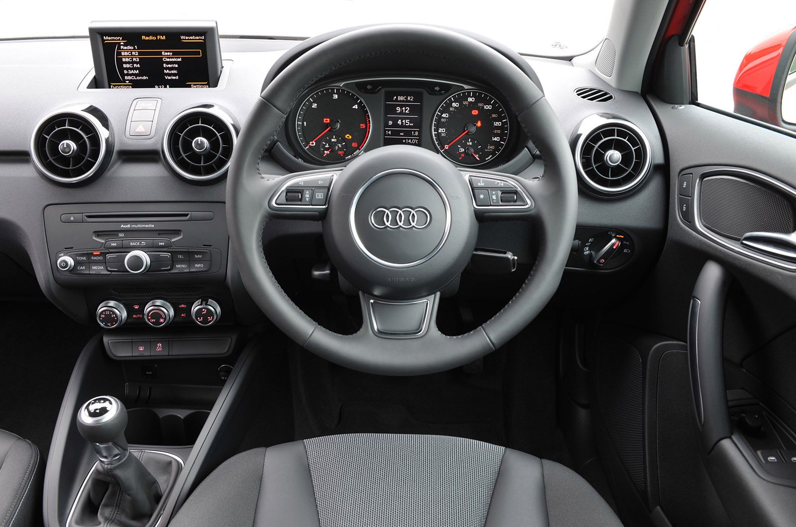 Car interior with a central infotainment display, steering wheel with controls, dashboard dials, and a manual gear stick.