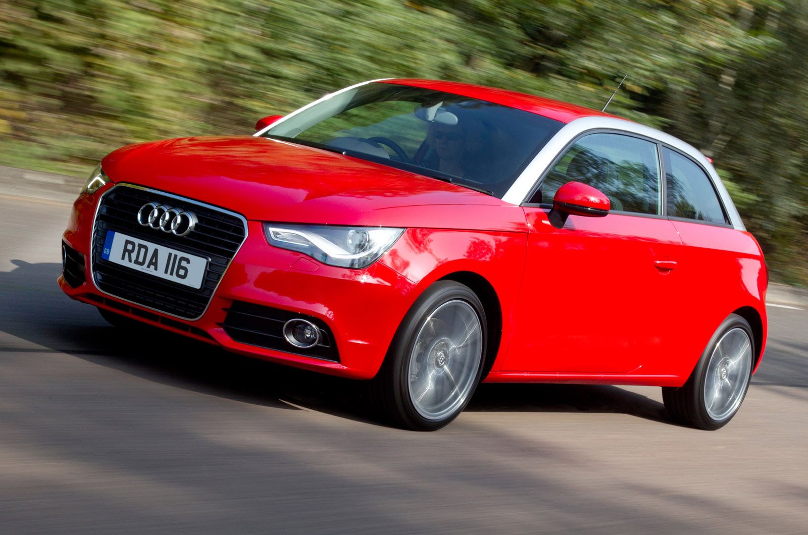 A red Audi car driving on a road, with trees blurred in the background, suggesting motion. License plate visible.