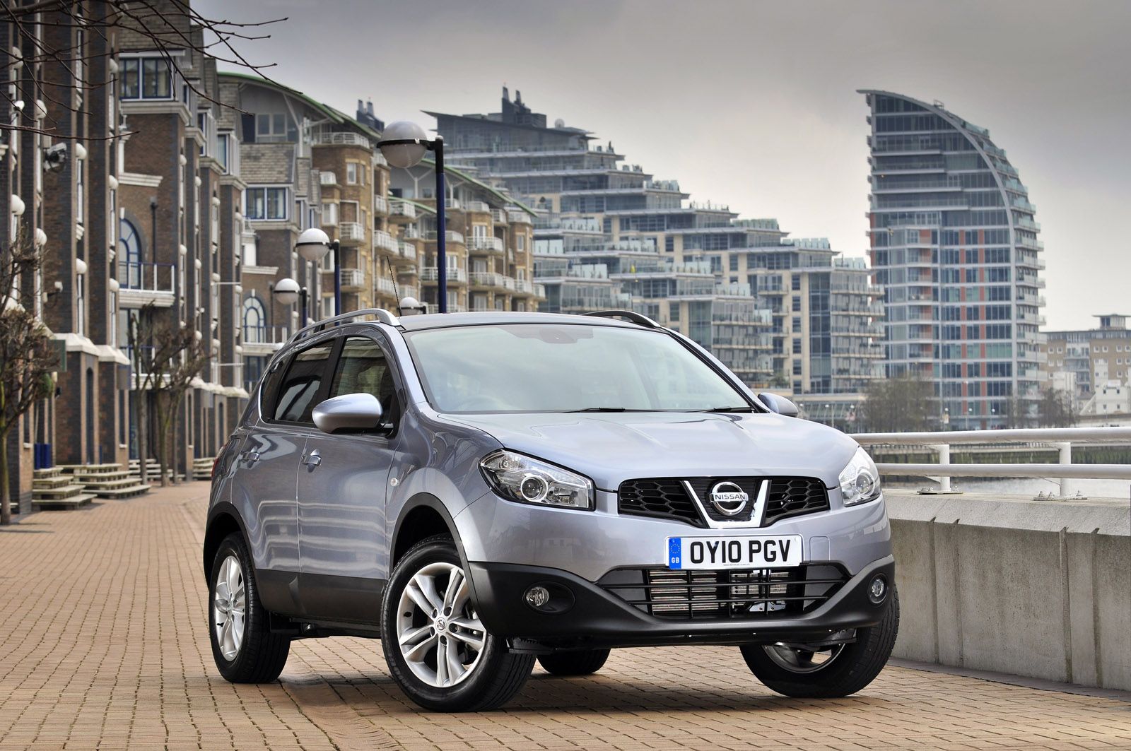 Silver Nissan Qashqai parked on a brick road in an urban area with modern buildings in the background under a cloudy sky.