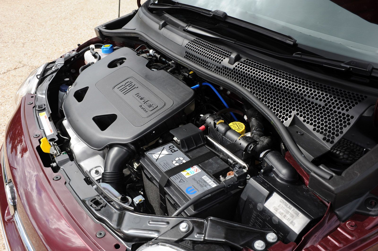 Car engine bay with a battery and an engine cover marked "MultiJet." The vehicle is maroon-colored.
