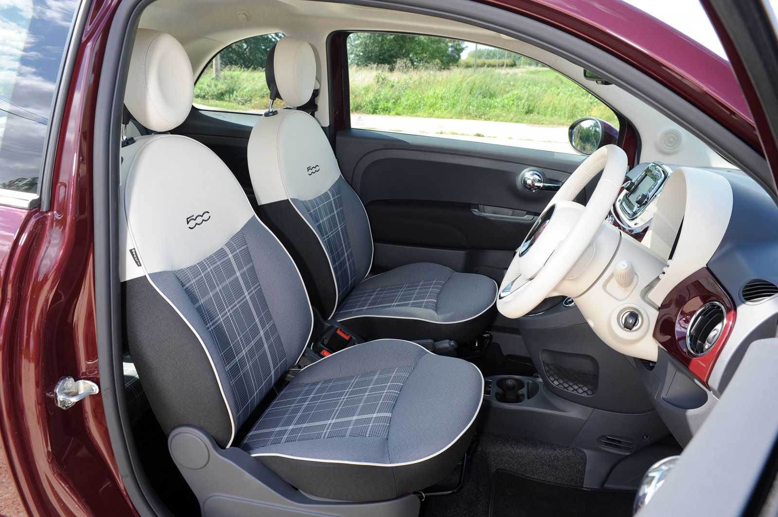 Interior of a small car with gray plaid seats, white and gray dashboard, and a white steering wheel. The door is open, showing lush greenery outside.