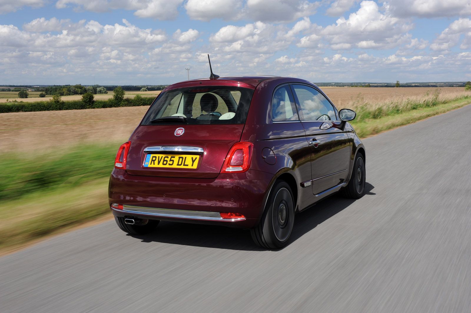 A maroon compact car with a license plate "RV65 DLY" drives down a rural road flanked by fields under a partly cloudy sky.