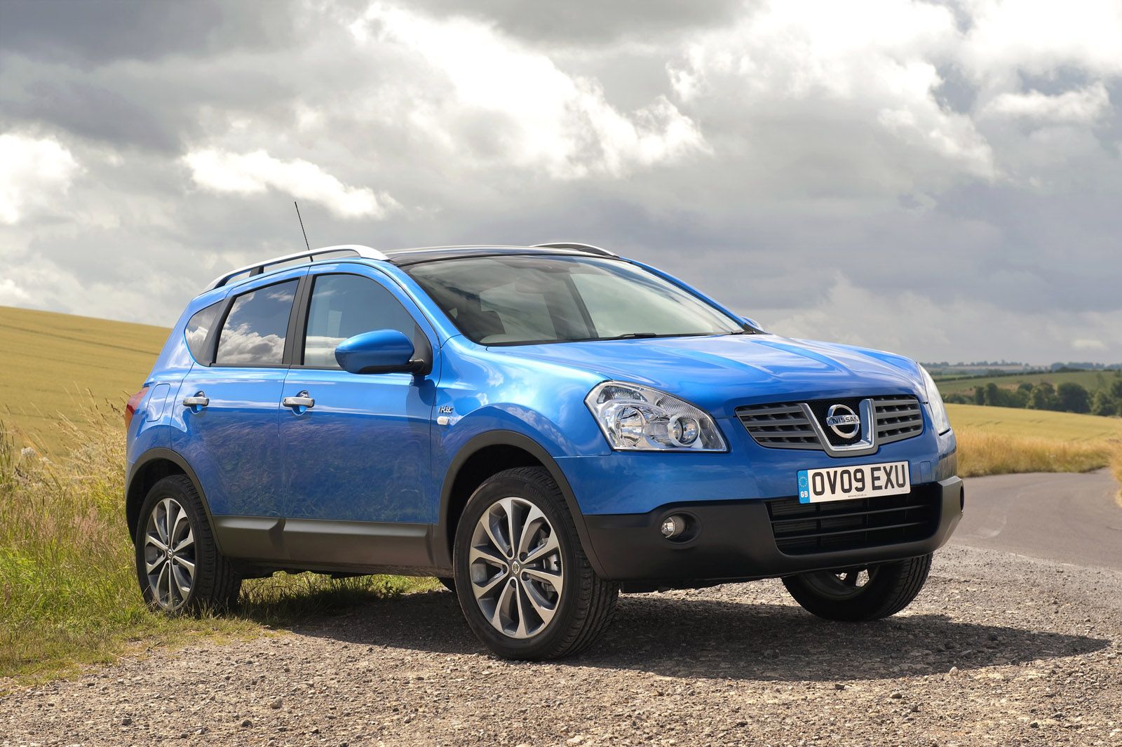 Blue SUV parked on a gravel path beside a field under a cloudy sky.