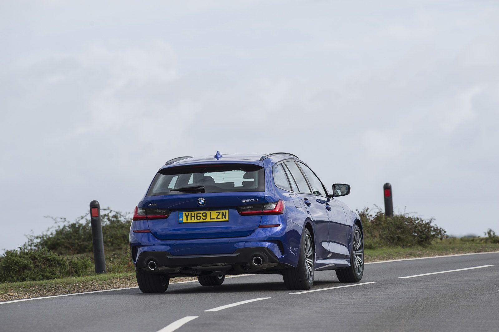Blue car driving on a winding road with cloudy skies, rear view showing license plate and dual exhaust.