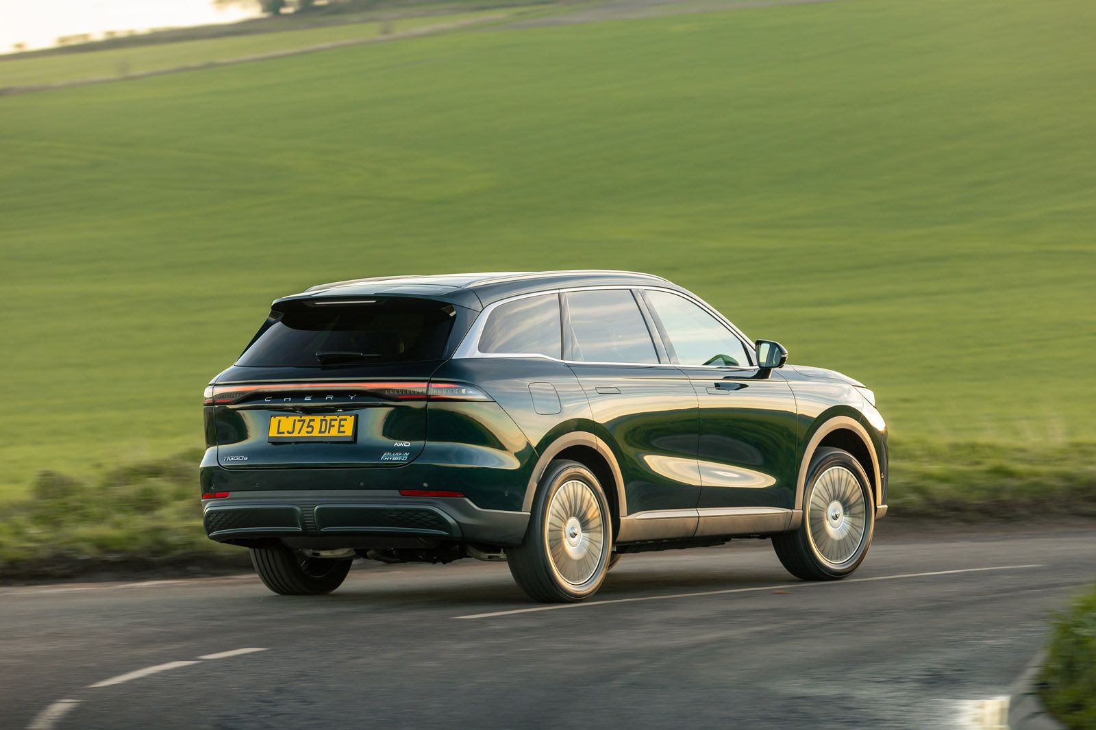A dark SUV driving on a country road with green fields in the background during daylight.