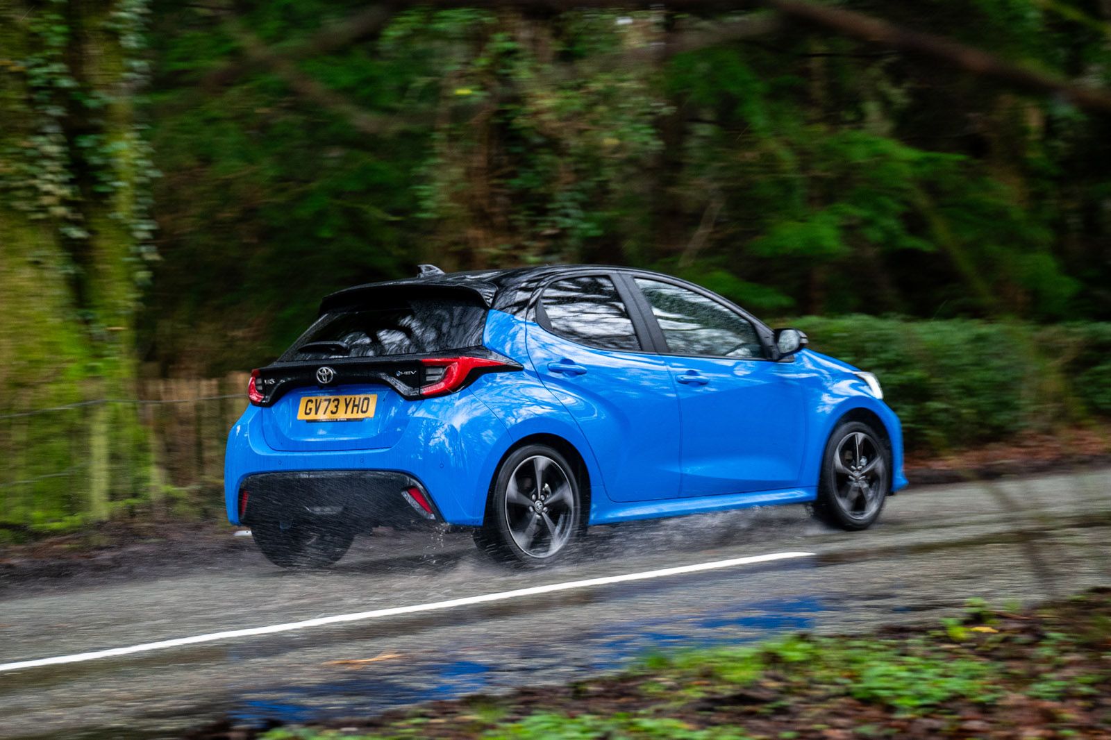 A blue car drives on a wet road through a forest, with blurred motion indicating speed.