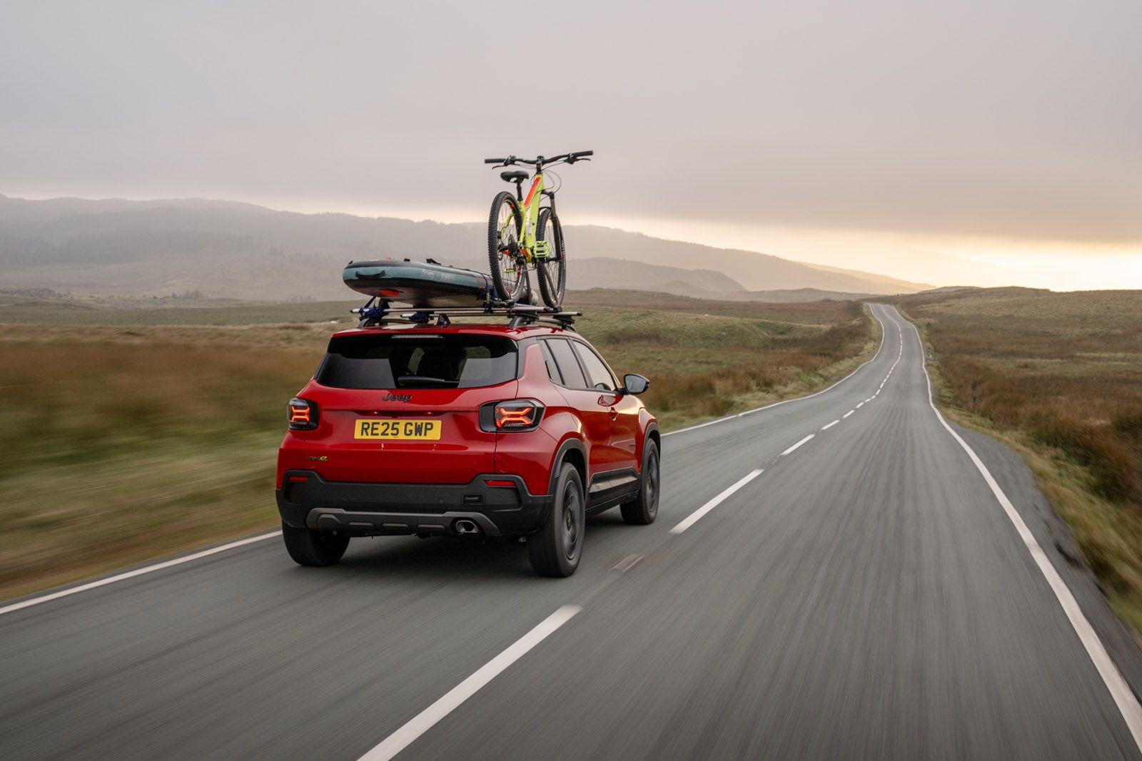 Red SUV with a bicycle and kayak on the roof drives down a deserted, winding road through a scenic, hilly landscape at dawn.