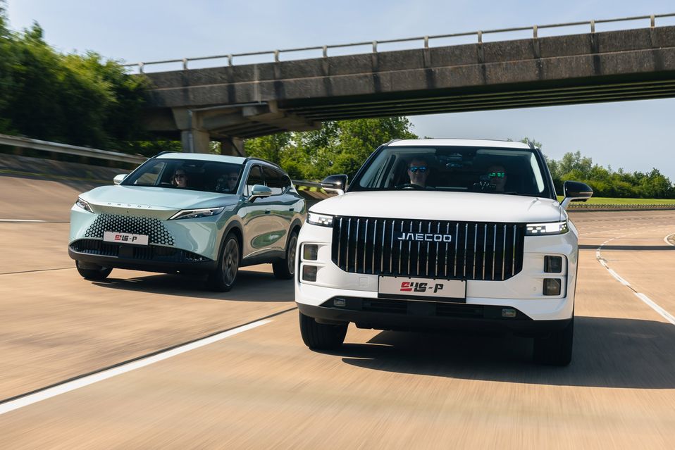 Two SUVs driving side by side on a sunny road under a bridge, with trees and greenery in the background.