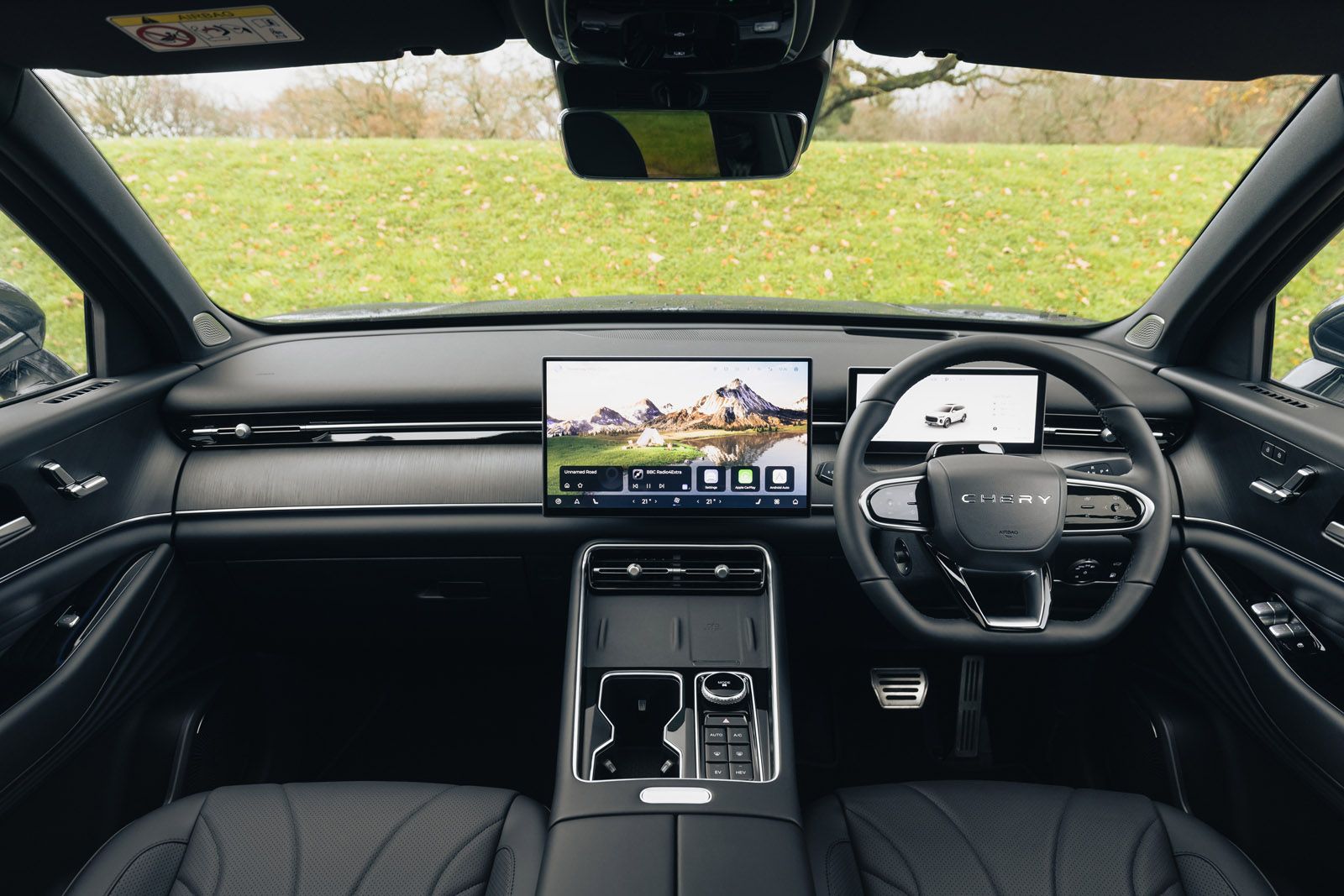 Modern car interior with a sleek dashboard, digital displays, and a black leather steering wheel, overlooking a grassy outdoor scene.