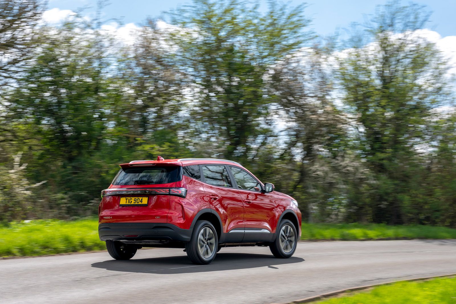 Red SUV driving on a curvy road with blurred trees in the background, under a partly cloudy sky.