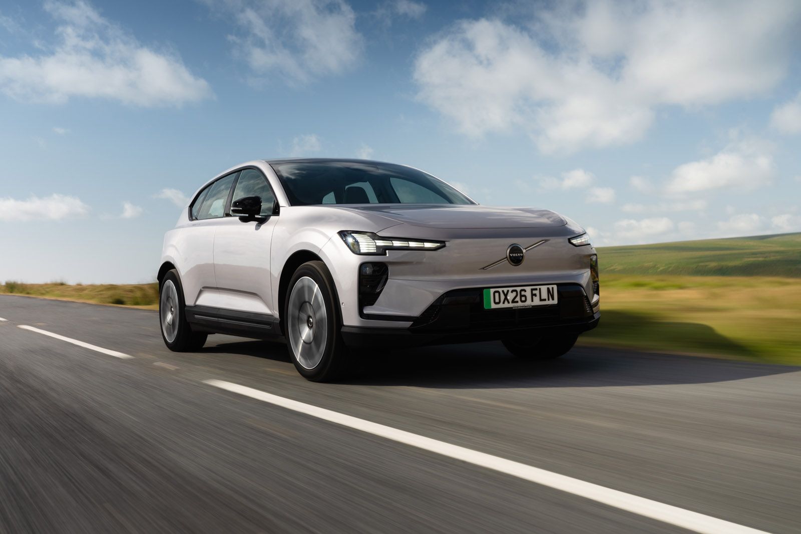 Silver SUV with sleek design driving on a country road under a blue sky with scattered clouds.