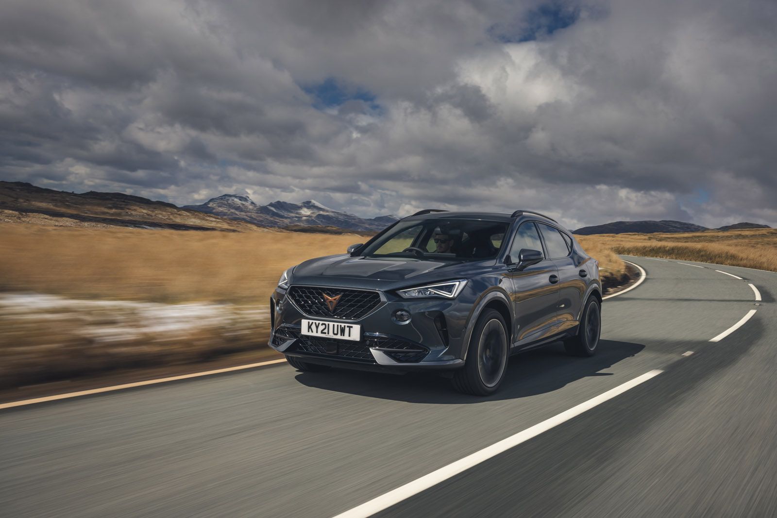 A sleek, modern SUV driving on a winding road through a scenic landscape with mountains and cloudy skies in the background.