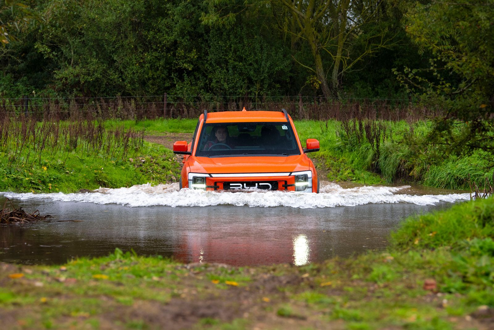 An orange SUV crossing a shallow river surrounded by lush greenery.