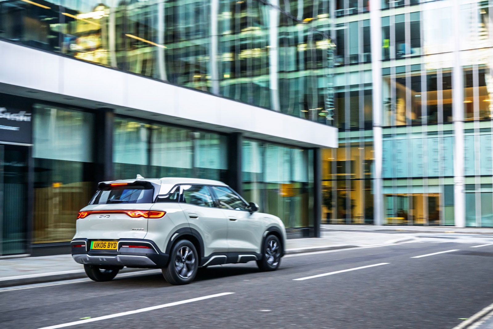 A white SUV drives down an empty city street with modern glass buildings reflecting light in the background.