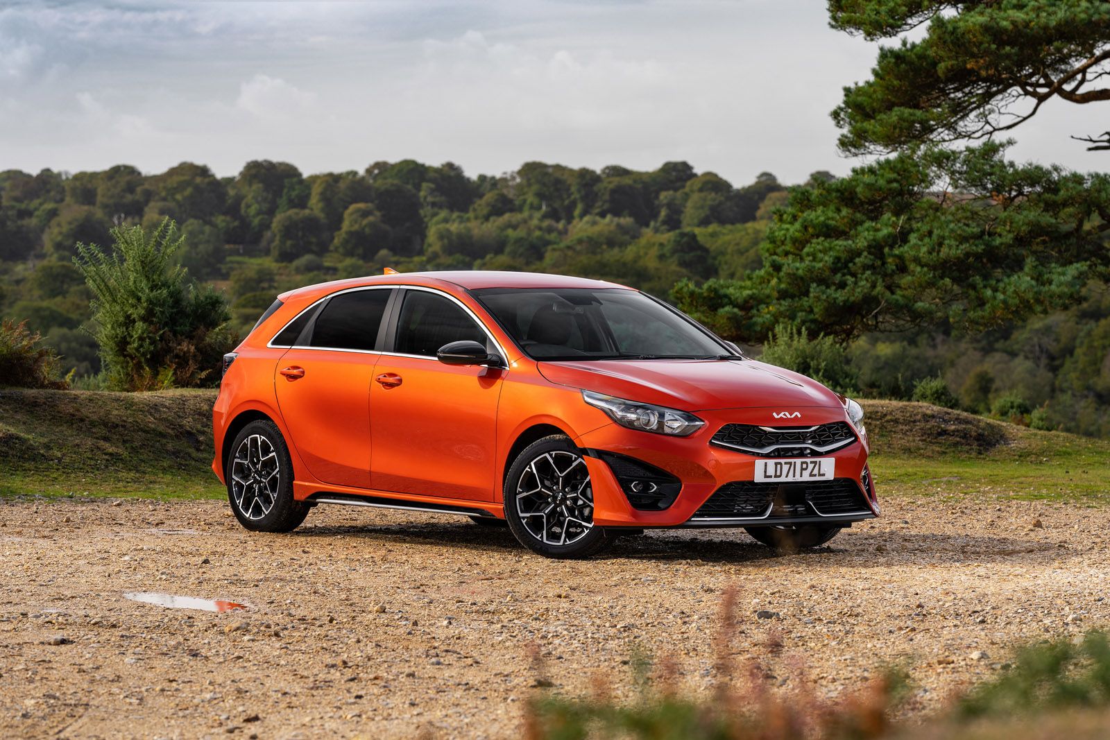 Orange Kia hatchback parked on a gravel path with a scenic forested background under a cloudy sky.