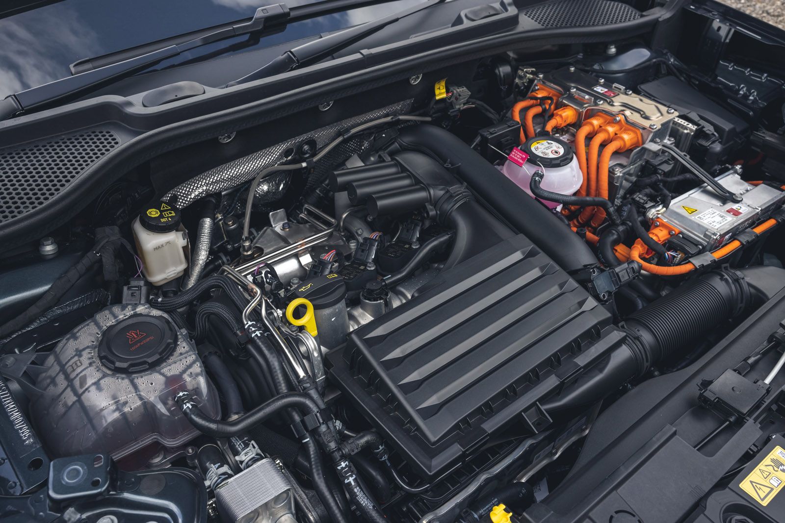 Close-up of a car engine bay featuring various components, including wiring, fluid reservoirs, and a distinctive orange high-voltage cable.