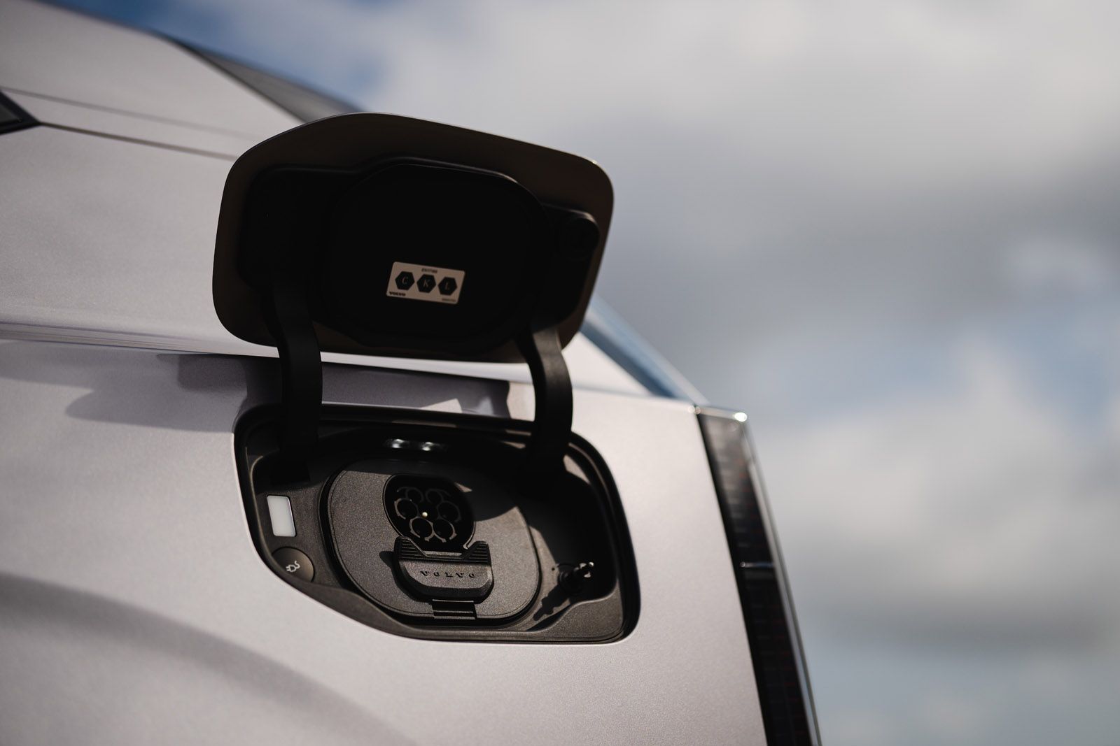 Close-up of an electric vehicle's charging port with an open cover, against a cloudy sky backdrop.