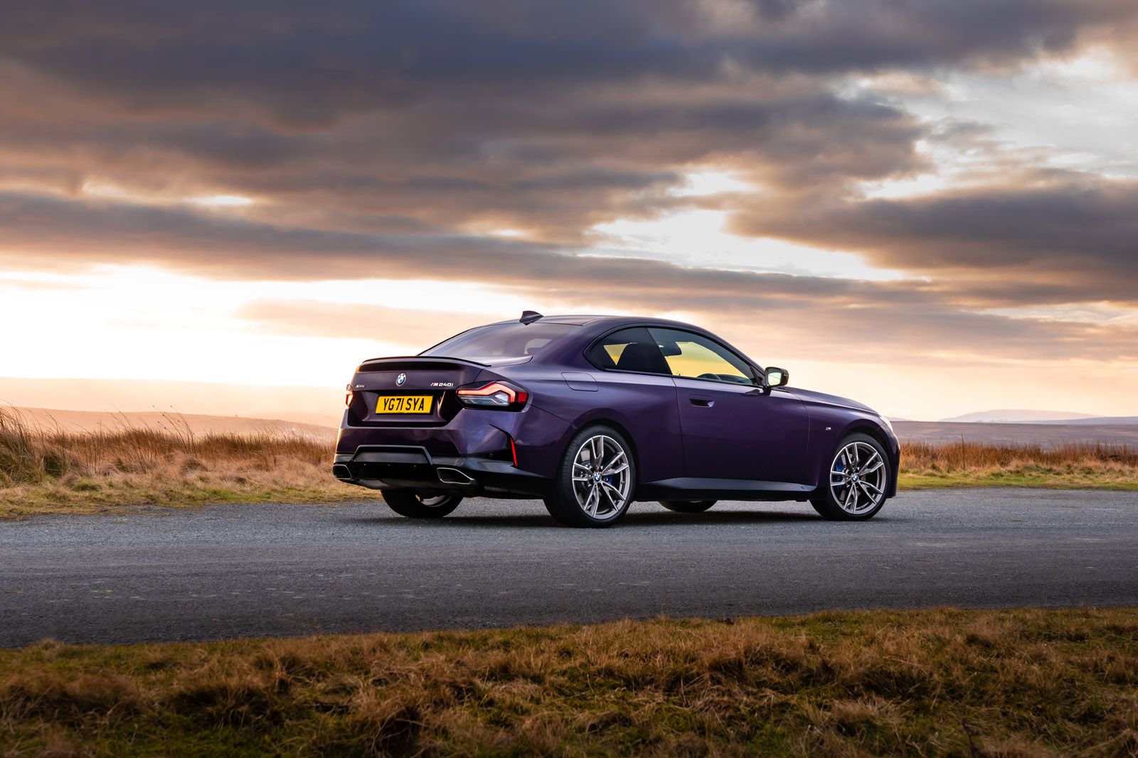 A sleek purple sports car parked on a rural road at sunset, with a dramatic cloudy sky and open fields in the background.