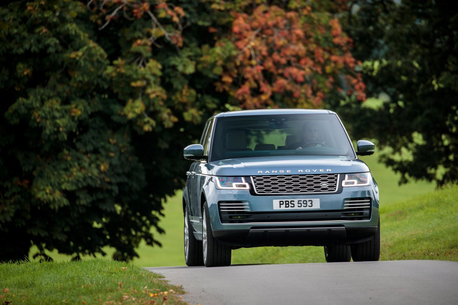 A blue Range Rover drives on a narrow country road, flanked by green trees with some autumn foliage.